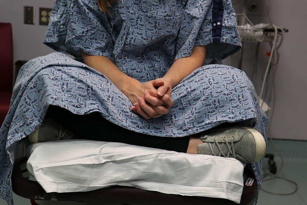 照片中，一个身份不明的病人的下半身穿着病号服，坐在病房的桌子上，等待一名性侵犯检查护士的检查。